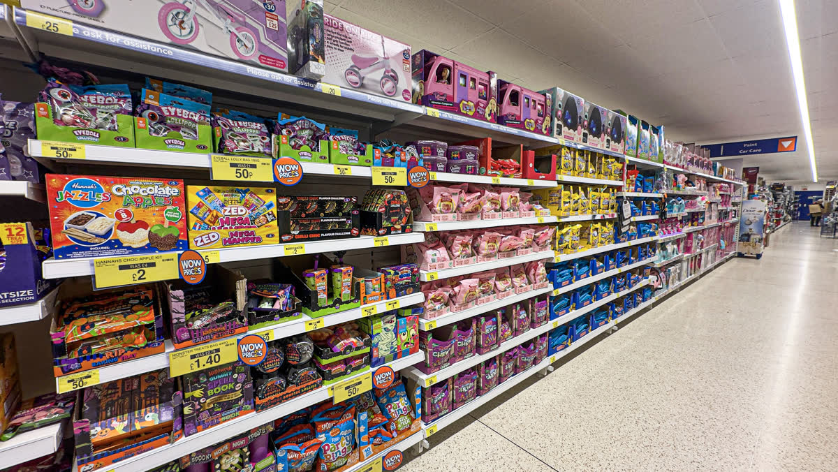 Interior view of toy & confectionary aisle at B&M discount store, 134-142 High Street, Gillingham, Kent, England, showing promotional displays, board games, puzzles, dolls and toy vehicles with yellow price labels — 스위치2판 가격, 얼마일까
